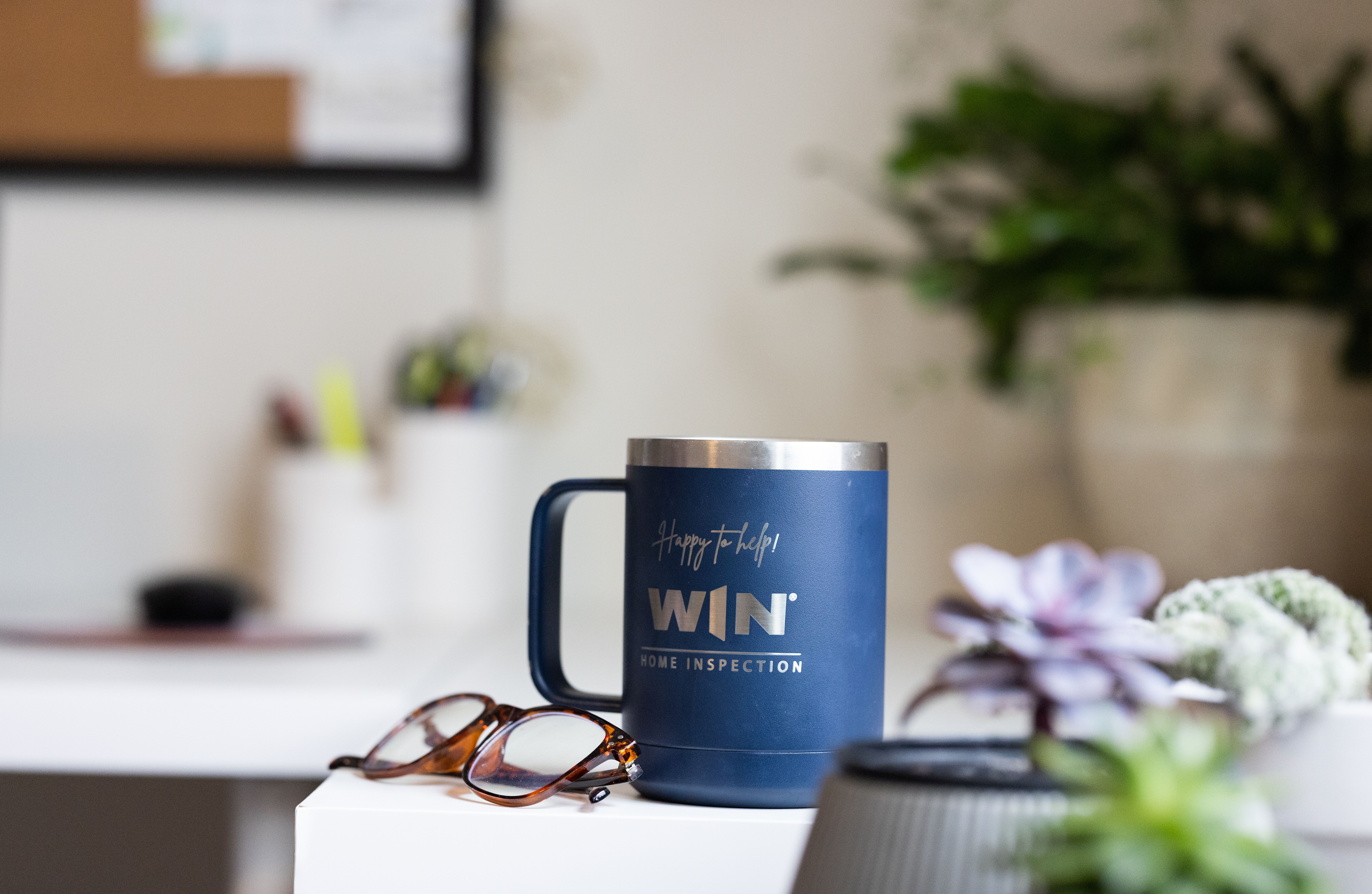 A blue “WIN Home Inspection” mug sits on a white desk beside eyeglasses, with blurred office supplies and plants in the background.